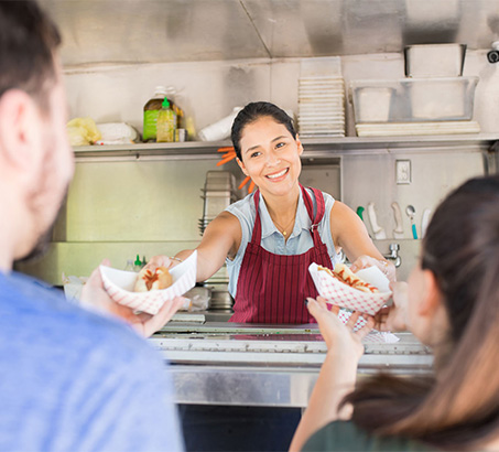 A woman serving two customers hotdogs from inside a food truck
